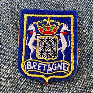 May include: A blue and yellow embroidered patch with a coat of arms featuring two white stylized animals, a crown, and the text "BRETAGNE".
