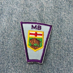May include: A purple, white, and yellow embroidered patch with the letters "MB" and a bison inside a shield. The patch is on a blue denim background.