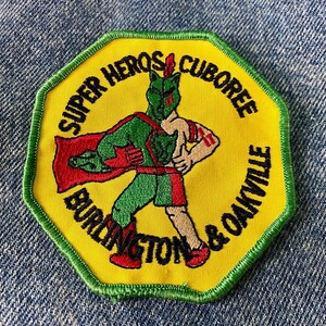May include: A yellow octagon-shaped patch with green stitching around the edges. The patch features a cartoon superhero with a red cape, green pants, and a red and white headband. The text on the patch reads "Super Heroes Cuboree Burlington & Oakville".