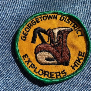 May include: A yellow and green embroidered patch with the text "Georgetown District Explorers Hike" and a cartoon image of a hiker with a backpack.