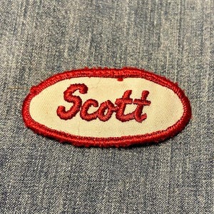 May include: An embroidered patch featuring the name "Scott" in red script. The patch has a white background and a red oval border. It is set against a blue denim fabric background.