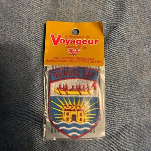 May include: A Thunder Bay patch from the World Wide Collectors Series. The patch features a shield design with the text "THUNDER BAY" at the top. The patch has a blue, red, yellow, and white colour scheme. The patch is in a clear plastic package with a yellow header.