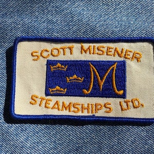 May include: A white embroidered patch with blue trim. The patch features the text "SCOTT MISENER STEAMSHIPS LTD." in gold lettering. A blue rectangle with three gold crowns and a gold letter "M" is in the centre of the patch.