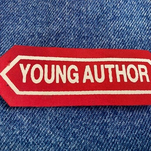 May include: Red fabric label with white text that reads "YOUNG AUTHOR" on a blue denim background.