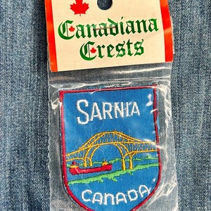 May include: A blue and red embroidered patch featuring the text "Sarnia, Canada" and a yellow bridge over a body of water. The patch is packaged in a clear plastic bag with a white card that reads "Canadiana Crests" with a red maple leaf.
