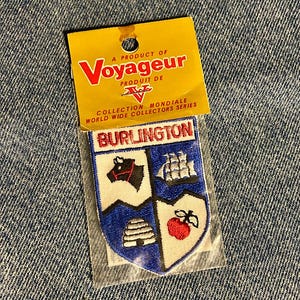 May include: A rectangular embroidered patch featuring a shield divided into four quadrants with a bull, a ship, a beehive, and an apple. The patch is labelled BURLINGTON and is attached to a yellow card marked A PRODUCT OF Voyageur.