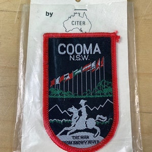 May include: A red, white, and blue woven patch with the text "COOMA N.S.W." and a depiction of a man on horseback. The patch is labeled "The Man from Snowy River" and is packaged in a clear plastic bag with a white card that reads "WOVEN BADGES by CITER".