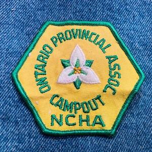 May include: A yellow and green embroidered patch with the text "Ontario Provincial Assoc Campout NCHA" and a white flower in the center.