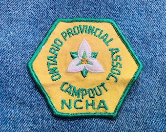 May include: A yellow and green embroidered patch with the text "Ontario Provincial Assoc Campout NCHA" and a white flower in the center.