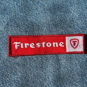 May include: A rectangular embroidered patch featuring a red background with the white text Firestone and a white square on the right side containing a red shield logo with a letter F inside. The patch is attached to a blue denim fabric surface.