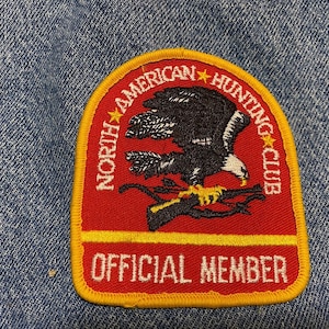 May include: Red and yellow embroidered patch with a white eagle and bow and arrow design. The text reads "North American Hunting Club" and "Official Member".