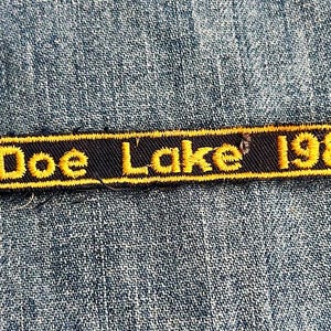 May include: Embroidered patch featuring the text "Doe Lake 1982" in yellow thread on a black background. The patch has a yellow border and a small green and white emblem. The patch is rectangular with banner-like ends.