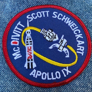 Peut inclure: Un écusson brodé bleu et blanc avec une bordure rouge. L'écusson représente une fusée, une orbite jaune et le texte "McDivitt Scott Schweickart Apollo IX".