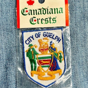 May include: A white embroidered patch with a blue border. The patch features the City of Guelph crest, which includes a green and white shield with a red and yellow stripe, a golden lion, and two figures in green and blue clothing. The text "City of Guelph" is written in a gold arc above the shield.