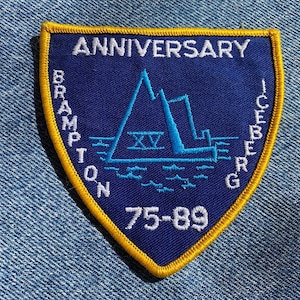 May include: Blue and white embroidered patch with a yellow border. The patch is shaped like a shield and features a stylized iceberg with the text "BRAMPTON ICEBERG" and "ANNIVERSARY 75-89" around the edge.