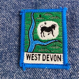 May include: A blue and green embroidered patch with a white background. The patch features a stylized image of a river and a cow with the text "WEST DEVON" below.