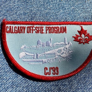 Peut inclure: Un écusson bleu clair avec une bordure rouge, portant l'inscription "CALGARY OFF-SITE PROGRAM" et "CJ'93". L'écusson présente une image brodée en blanc d'un paysage urbain et une feuille d'érable rouge.