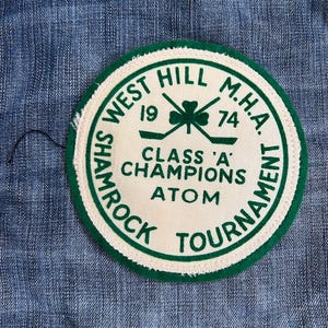 May include: A white and green circular patch with the text "West Hill M.H.A. Shamrock Tournament" and "Class 'A' Champions Atom" in green lettering. The patch also features a green shamrock with two crossed hockey sticks.