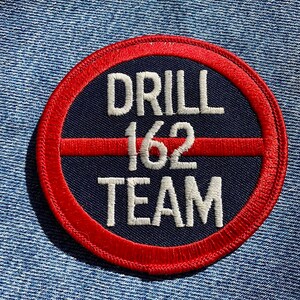 May include: Circular embroidered patch with a red border and a navy blue center. The patch features the words "DRILL 162 TEAM" in white, with a red horizontal line through the number 162. The patch is set against a denim background.