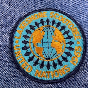 May include: A blue and yellow embroidered patch with the text "United Nations Lakeshore Scoutoree 1997" and a globe in the center.