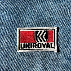 May include: A rectangular embroidered patch featuring the Uniroyal logo. The design includes a white border, a red background, and a black stylised letter K graphic. The word UNIROYAL is stitched in black capital letters on a white base.