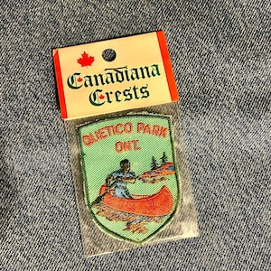 May include: A shield-shaped embroidered patch featuring a person paddling an orange canoe on light green water. The text reads QUETICO PARK ONT. The patch is inside a clear plastic sleeve attached to a white header card labelled Canadiana Crests.