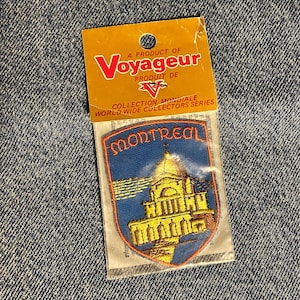 May include: A souvenir embroidered patch featuring a blue background with the word MONTREAL in orange letters above a yellow building illustration. The patch is inside a clear plastic sleeve attached to a yellow cardboard header card with the text Voyageur.