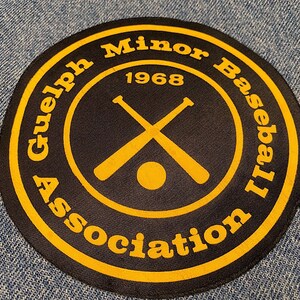 May include: A black and yellow circular patch with the text "Guelph Minor Baseball Association 1968" around the edge. The center of the patch features two crossed baseball bats and a baseball.