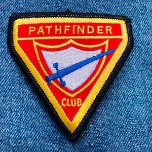 May include: A triangular patch with a yellow border and red background. The patch features a white shield with a blue sword and the text "PATHFINDER CLUB".