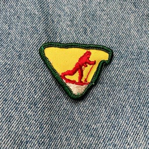 May include: A green, yellow, and white triangular patch with a red skier in the center. The patch is sewn onto a blue denim fabric.