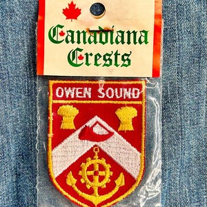 May include: A red and white embroidered patch with the text "Owen Sound" in yellow lettering. The patch is in a plastic bag with a white label that says "Canadiana Crests" in green and red lettering.