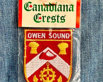 May include: A red and white embroidered patch with the text "Owen Sound" in yellow lettering. The patch is in a plastic bag with a white label that says "Canadiana Crests" in green and red lettering.