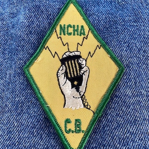 May include: A yellow diamond-shaped patch with a green border. The patch features a white hand holding a black microphone with a green cord. The text "NCHA C.B." is embroidered in green on the patch.