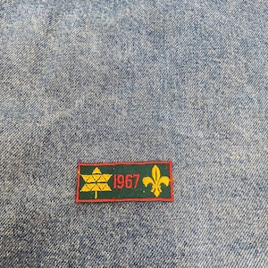 Peut inclure: Un écusson brodé vert et jaune avec un arbre stylisé, l'année "1967" et une fleur de lys.