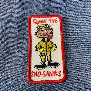 May include: Red, white and yellow embroidered patch featuring a cartoon character wearing goggles and a yellow jacket. The text "Damn the Sno-Snakes" is on the patch.