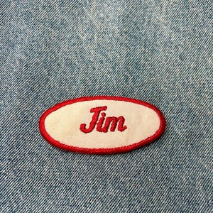 May include: A white oval name tag with a red border. The name "Jim" is embroidered in red on the tag.