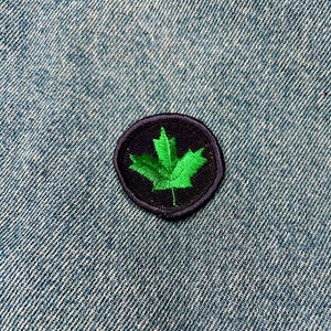 May include: A green maple leaf embroidered on a black circular patch. The patch is sewn onto a blue denim fabric.