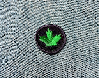 Puede incluir: Un parche circular negro con una hoja de arce verde bordada. El parche está cosido en una tela vaquera azul.