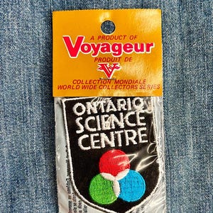 May include: A black and white embroidered patch with the text "Ontario Science Centre" in white lettering. The patch features a black shield with three overlapping circles in red, green, and blue.