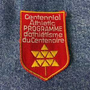 May include: Red and yellow embroidered patch with the text "Centennial Athletic Programme d'athlétisme du Centenaire" and a stylized yellow maple leaf.