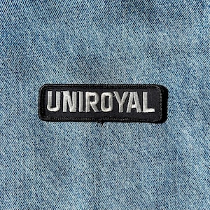 May include: A rectangular black embroidered patch featuring the white text UNIROYAL in a bold, sans-serif font. The patch has a black stitched border and is attached to a blue denim fabric background with a visible diagonal twill weave pattern.