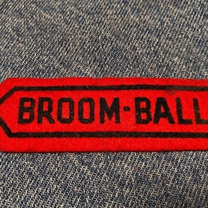 May include: Red felt patch with black outline and the text "BROOM-BALL" in black.