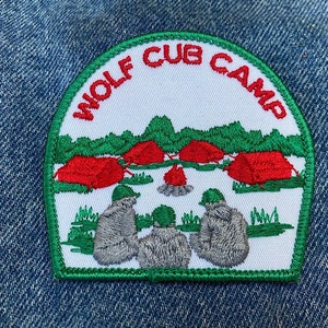 May include: A white embroidered patch with green trim, depicting a camping scene with red tents, a campfire, and three figures. The text "Wolf Cub Camp" is written in a red arc around the scene.