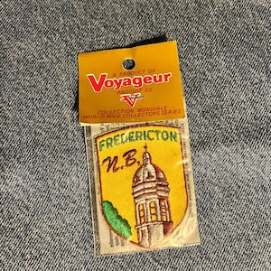 May include: A Voyageur brand embroidered patch in a clear plastic sleeve with a yellow header card. The patch features a yellow shield shape with green text reading FREDERICTON N.B. and an illustration of a domed building in red and white.