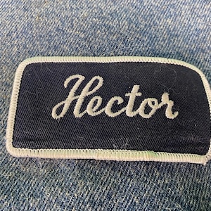 Peut inclure: Un patch en tissu bleu marine avec des coutures blanches autour des bords. Le mot "Hector" est brodé en blanc sur le patch.