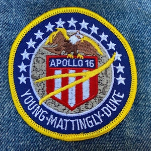 Peut inclure: Un écusson brodé bleu et blanc avec une bordure jaune. L'écusson représente un aigle chauve aux ailes déployées, un bouclier à rayures rouges et blanches, et le texte "Apollo 16 Young Mattingly-Duke".
