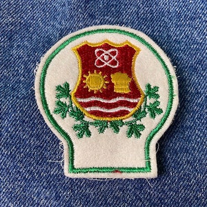 Peut inclure: Écusson en feutre blanc avec une bordure verte. L'écusson présente un bouclier rouge avec un motif blanc et jaune. Le bouclier est entouré de feuilles vertes.