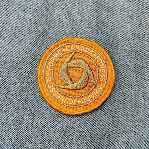 May include: A round, orange patch with white embroidered text that reads "En Forme Canada Fitness Award Jeux". The patch features a stylized gold-colored symbol of a person exercising.