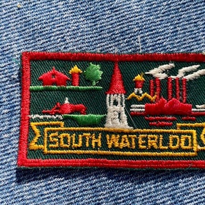 May include: A red, green, and white embroidered patch with the text "South Waterloo" in yellow. The patch features a small house, a tree, a tall building, and a factory with smoke stacks.