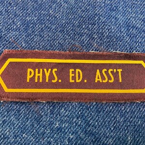 May include: A maroon and gold patch with the text "PHYS. ED. ASS'T" in yellow lettering. The patch has a gold border and is sewn onto a blue denim fabric.